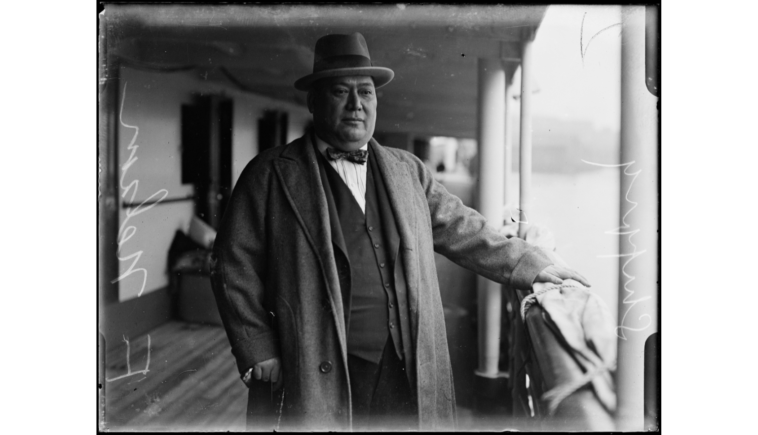 Well-dressed man in a hat, bowtie and coat standing on the deck of a ship looking at the view