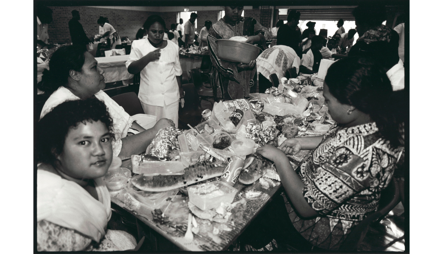 People sitting at and serving food on a long table covered in food containers and drink cans 