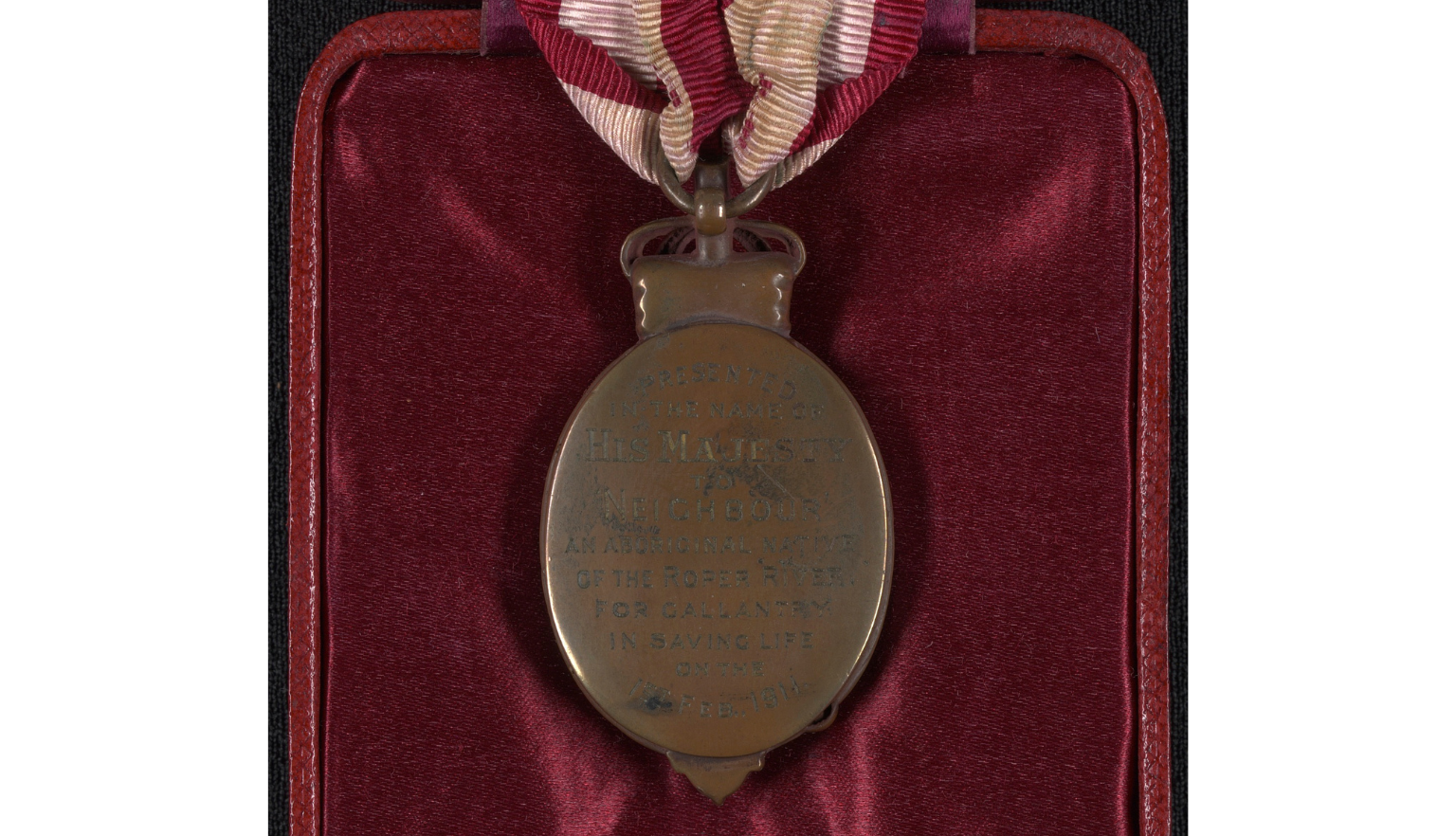 The reverse of an oval shaped bronze medal held by a red and white striped ribbon. On the back of the medal are the words 'Presented in the name of His Majesty to Neighbour, an Aboriginal native of the Roper River for gallantry in saving life on the 1st Feb 1911'