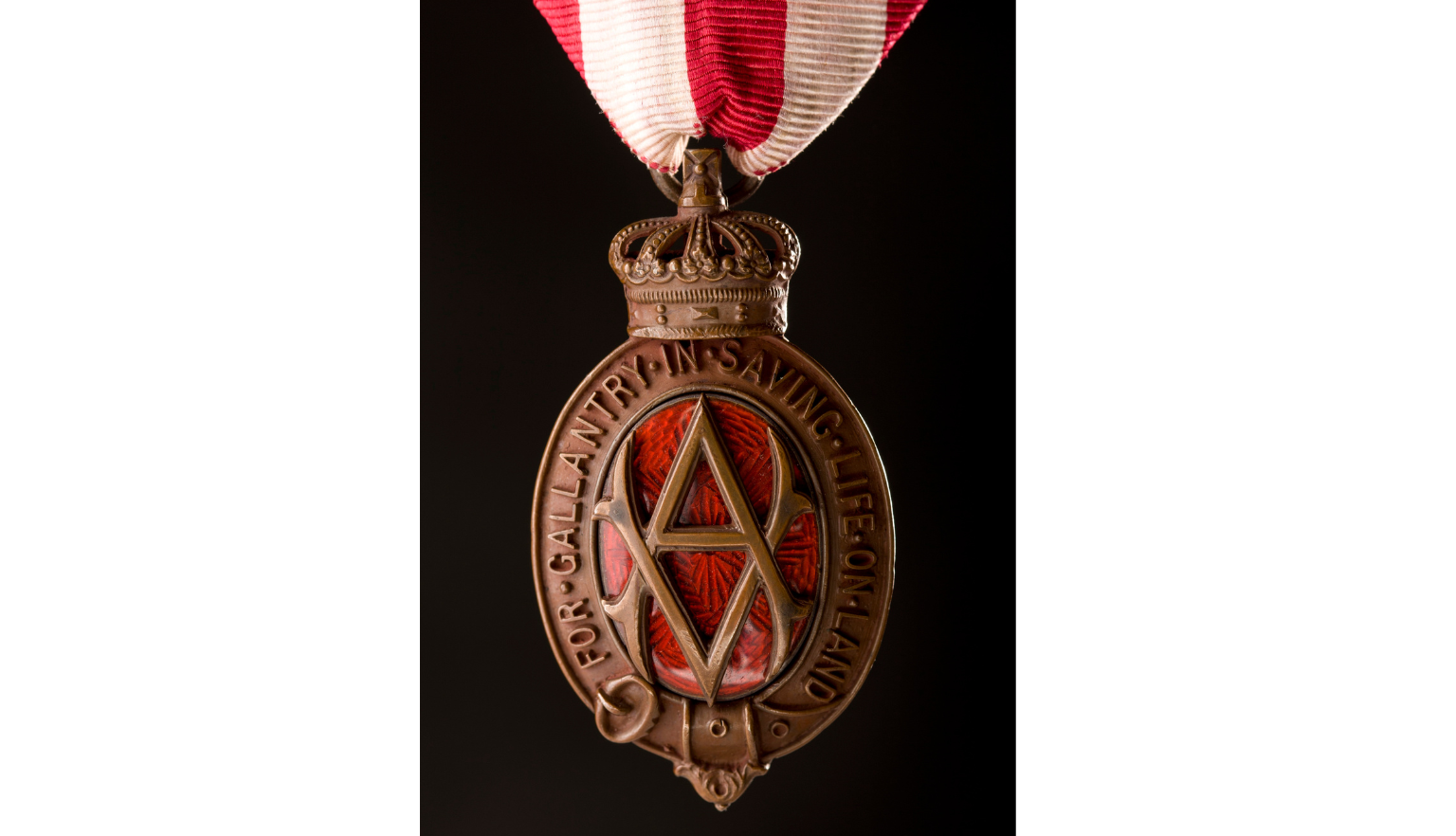 An oval shaped bronze medal. The oval is made of a belt buckled at the bottom. At the top is a stylised royal crown. In the centre of the oval is a monogram made up of an intertwined "V.A." Behind the monogram is pattered red glass. Around the oval in raised letters are the words 'For gallantry in saving life on land'. The medal is held by a ribbon of red and white vertical stripes 