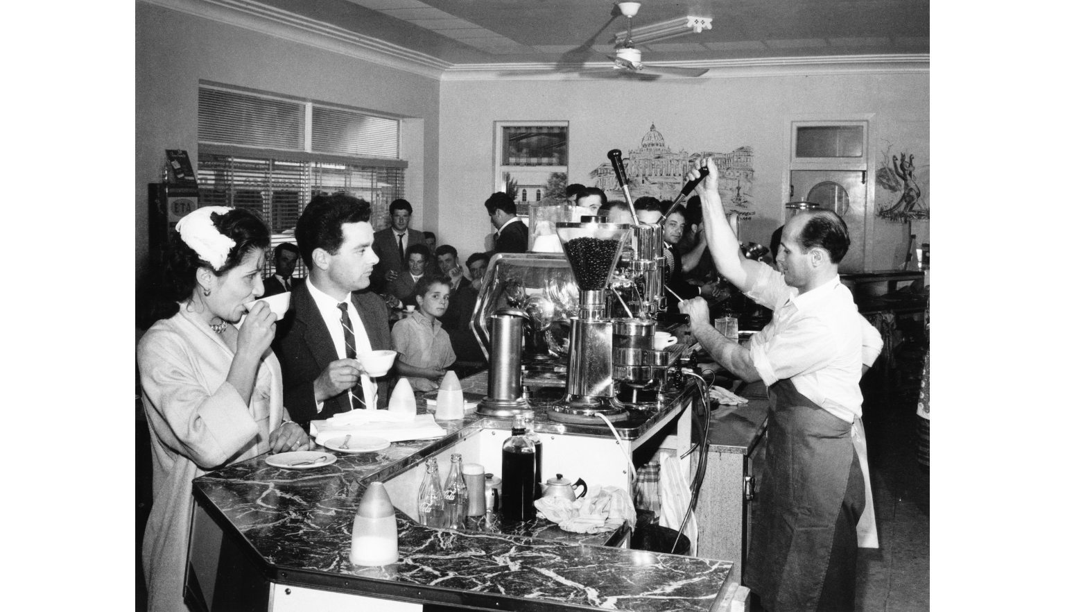 A black-and-white photograph of a busy café scene, with a man operating an espresso machine behind the counter. Customers stand or sit at the counter, drinking coffee.