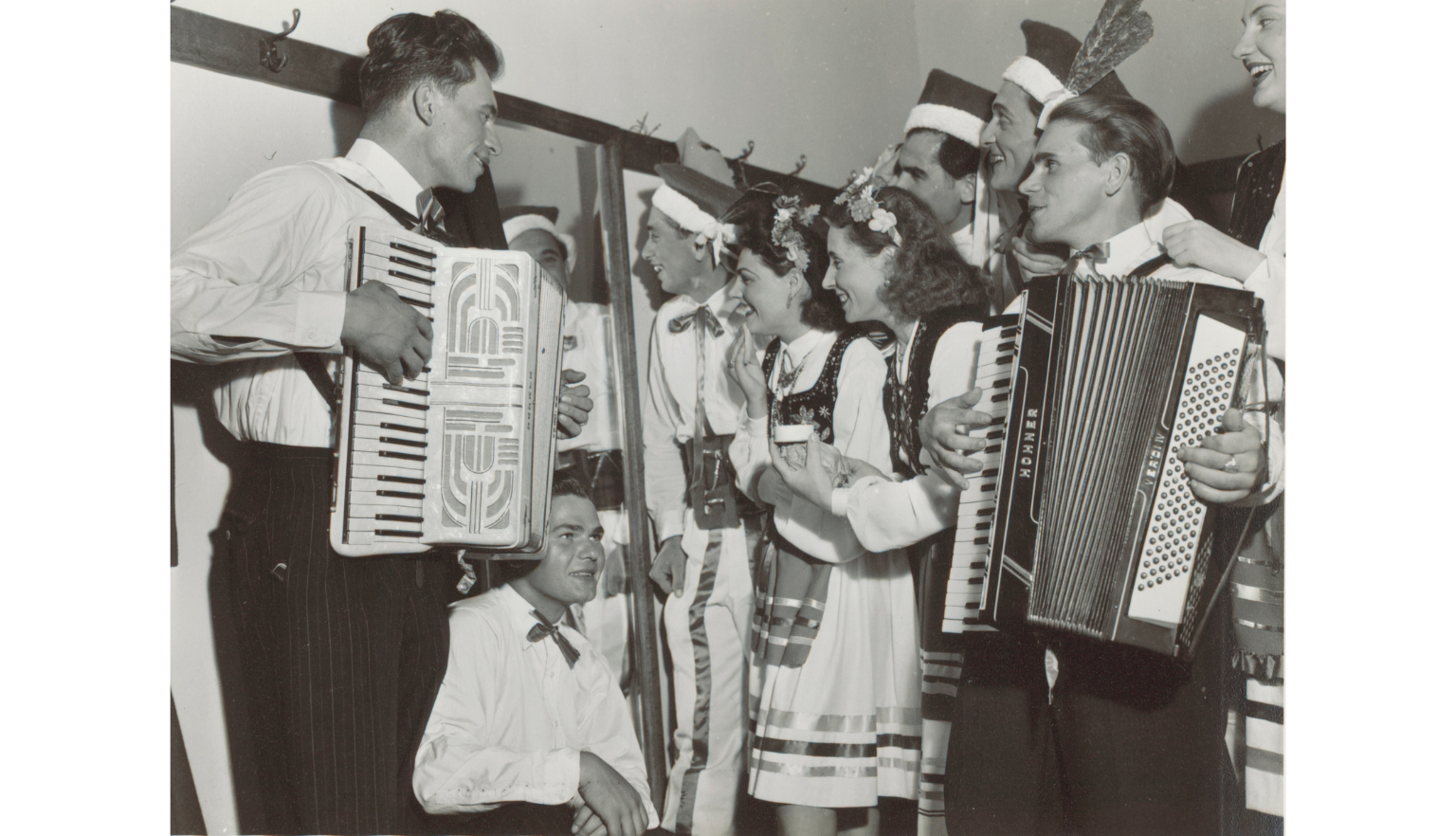 A black-and-white photograph of a group of people in traditional Polish folk costumes. Two men play accordions, while others stand and smile, some in conversation.