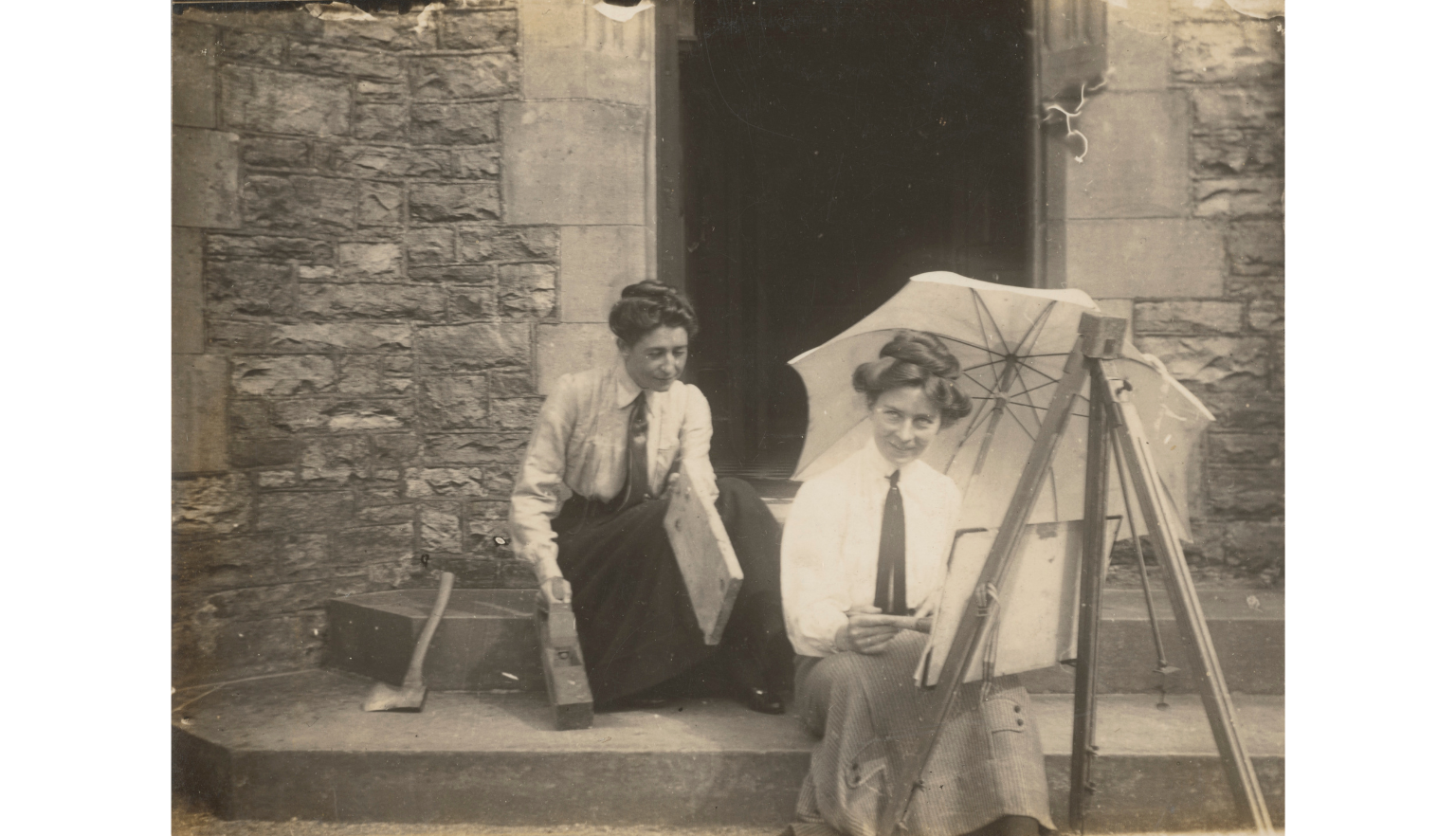 Sepia-toned photo of Eirene Mort and Nora Weston sitting the steps in front of a building with art supplies and an umbrella