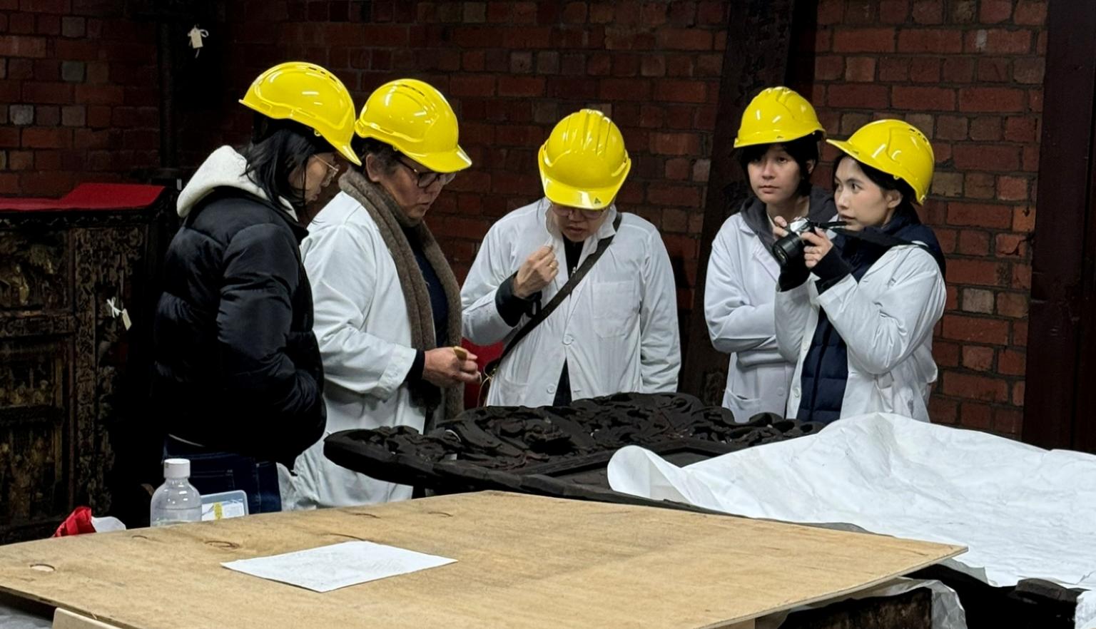 Five people wearing coats and hard hats are assembled around a table looking at a large item partially covered in cloth. 
