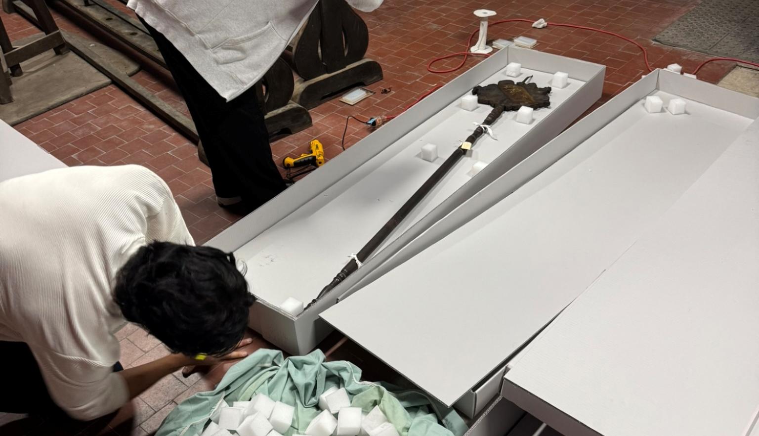 Two conservators stand on the left-hand side of this image. One is assembling a box and the other is photographing an item in the box. 