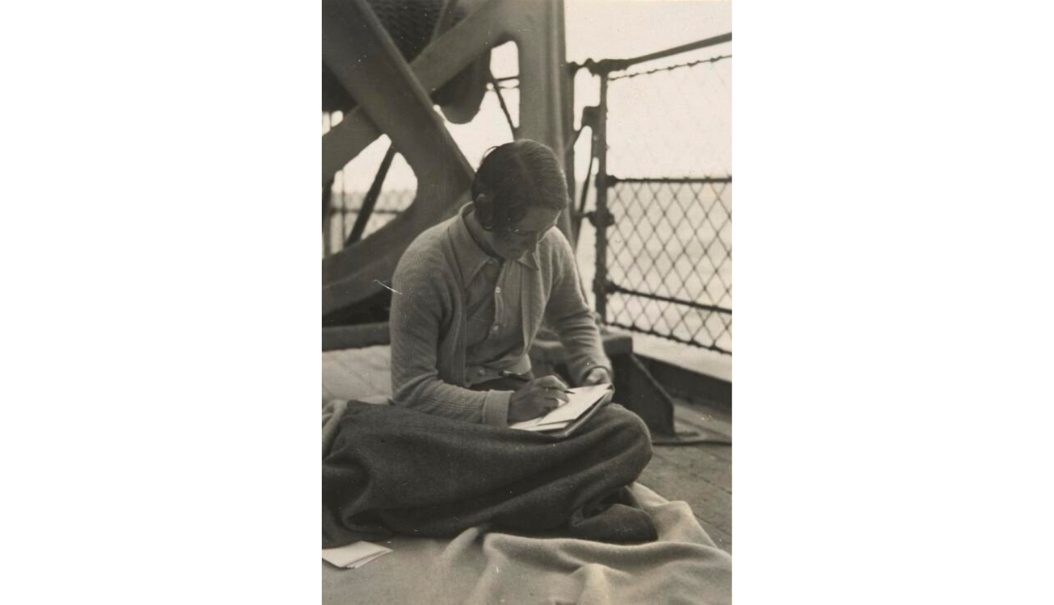 Black and white image, of a person sitting cross-legged writing, in the background is the railing of a boat deck the ocean beyond it