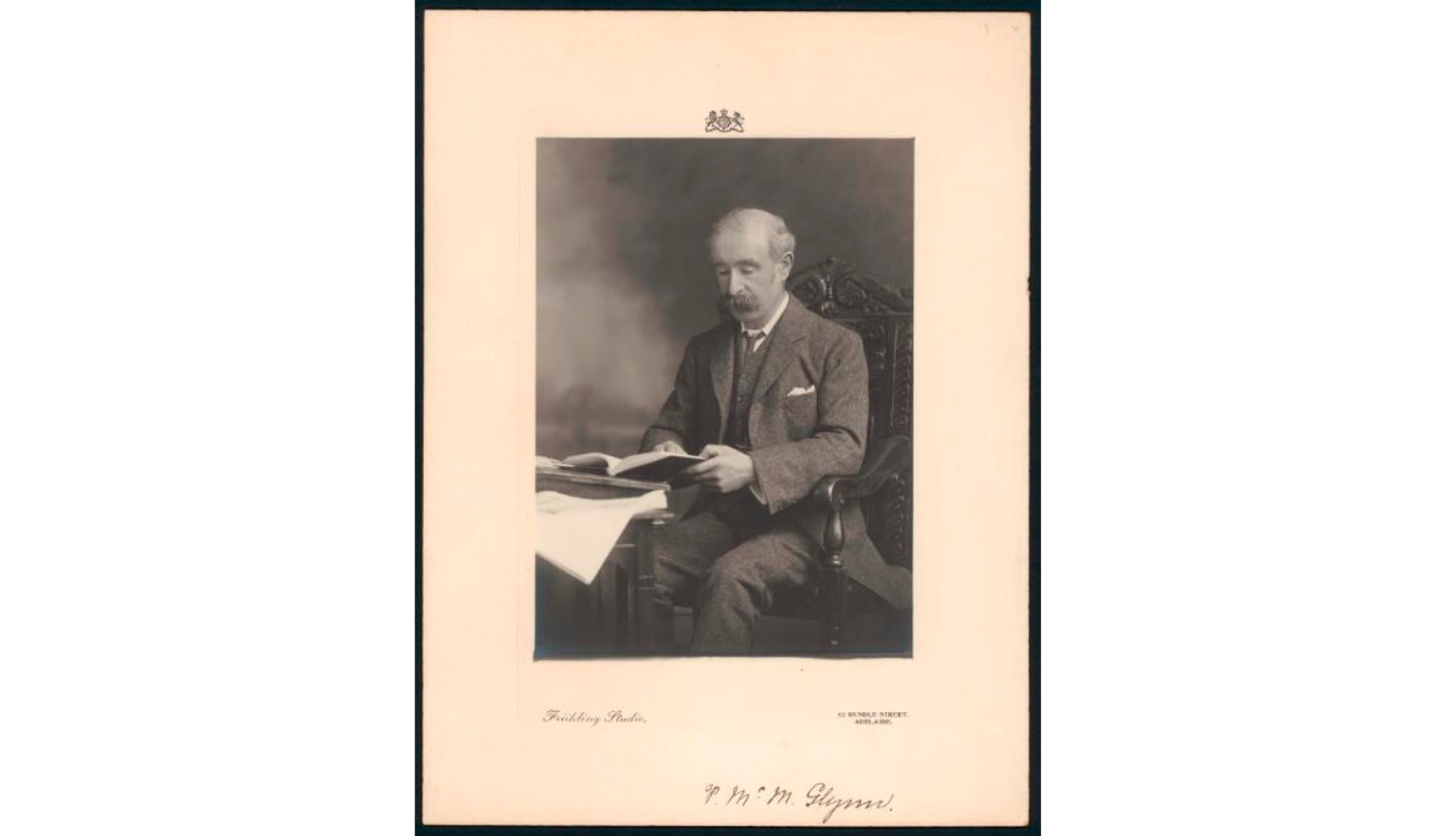 Black and white picture of Patrick McMahon Glynn seated at a table reading a book, picture is stuck to aged paper which bears his signature 