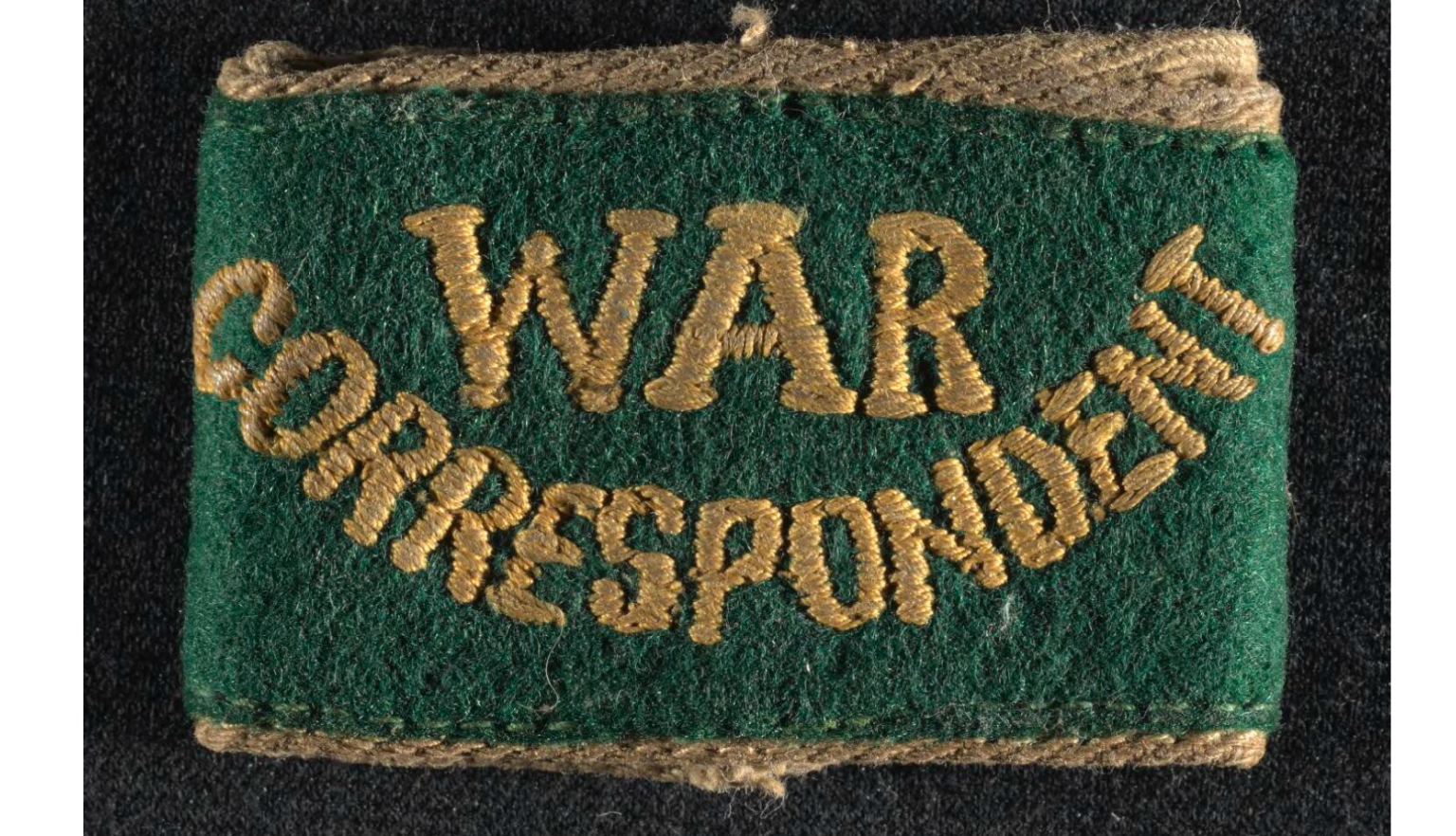 Photograph of green cloth badge, gold edging, 'War Correspondent' stitched in gold full caps.