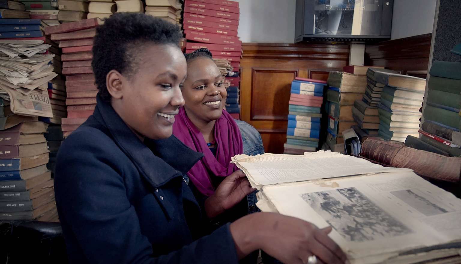 Two smiling women examining an open book with books piled high around them