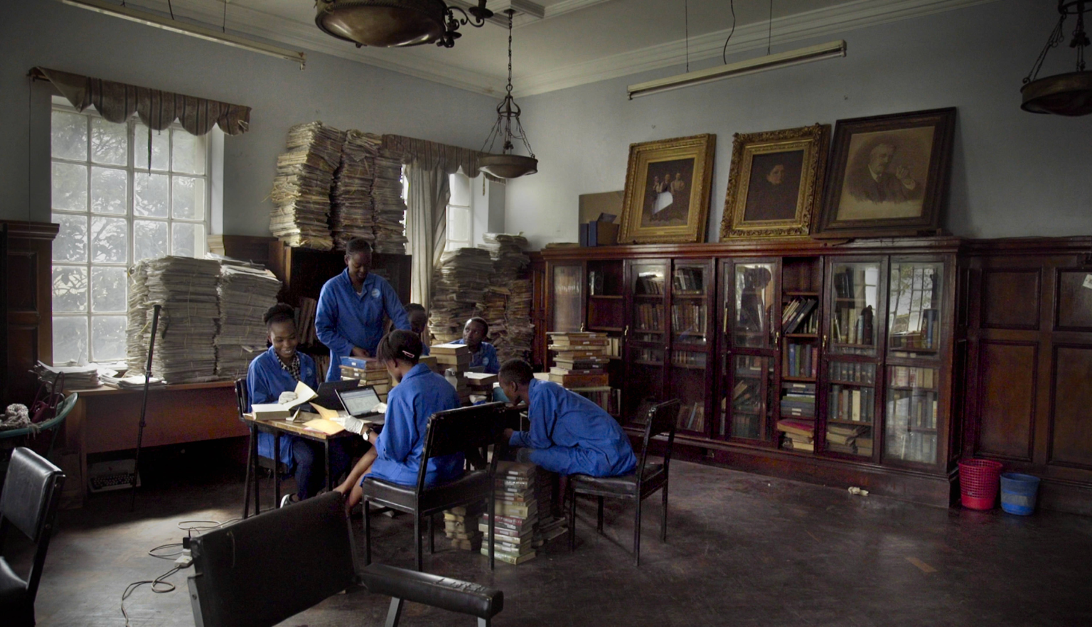5 people in blue grouped at a table in front them various open books, book stacks and laptops, the walls behind them stacked high with books and art atop of bookshelves