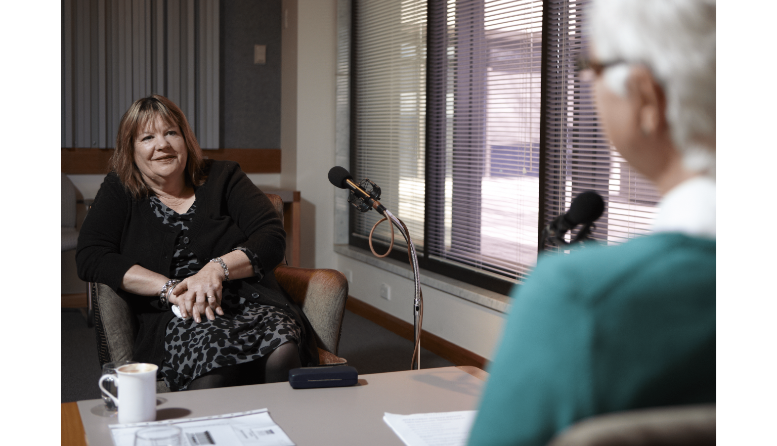 Woman sitting across from someone who is sitting out of frame, only visible by a shoulder in a teal sweater, in an interview format