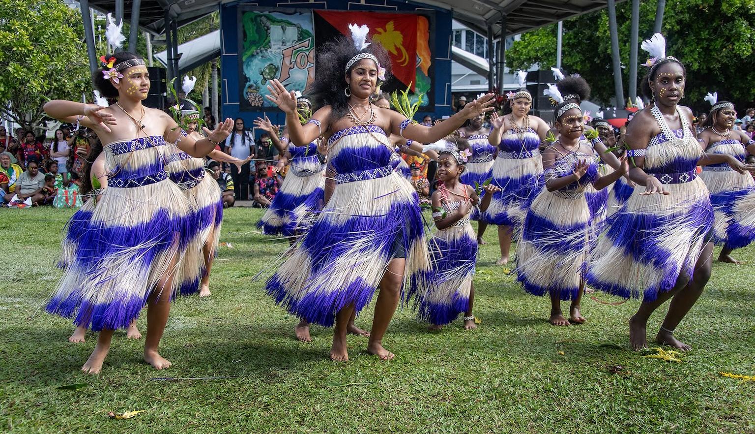 Several women dancing in rows with their arms out, wearing tassled purple and white dresses