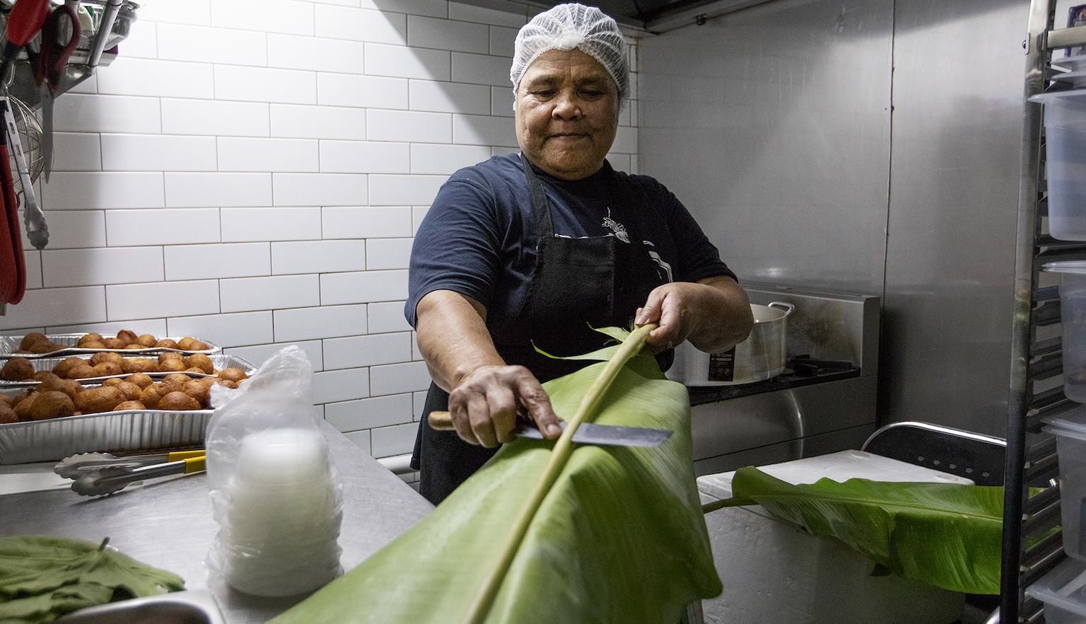 Woman in a hairnet running a knife along a banana leaf in a small kitched
