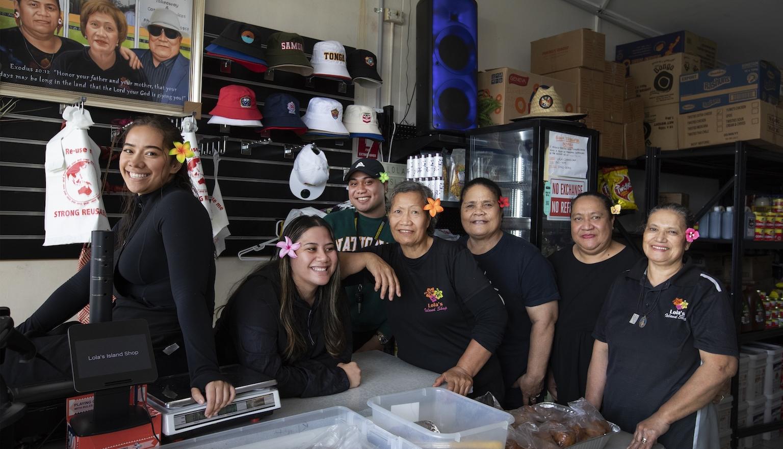 Women of various ages, some with flowers in their hair, standing behind the counter of a small grocery small smiling