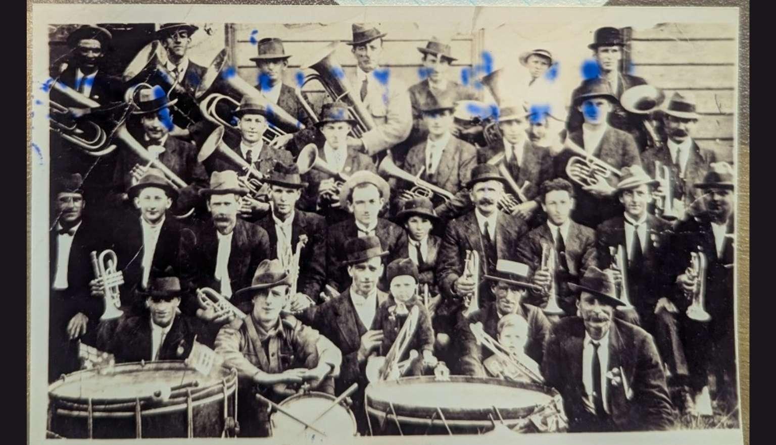 A vintage photograph featuring a large group of men dressed in early 20th-century attire, many wearing hats. They appear to be a marching band or musical group, holding various brass instruments.