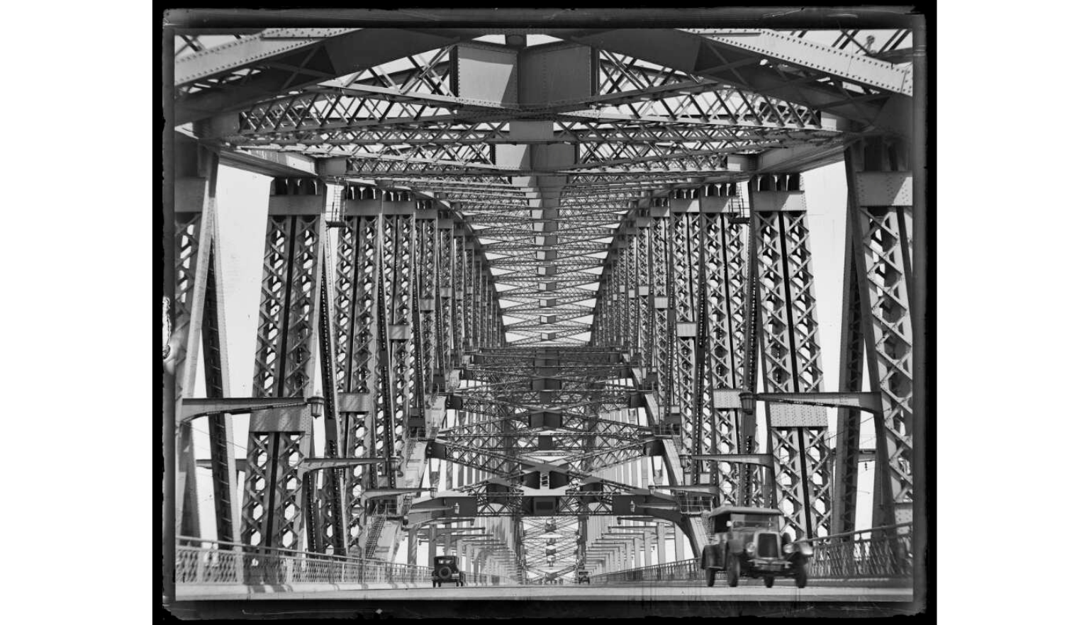 A black and white photograph showing the intricate steel truss structure of a large bridge extending into the distance.