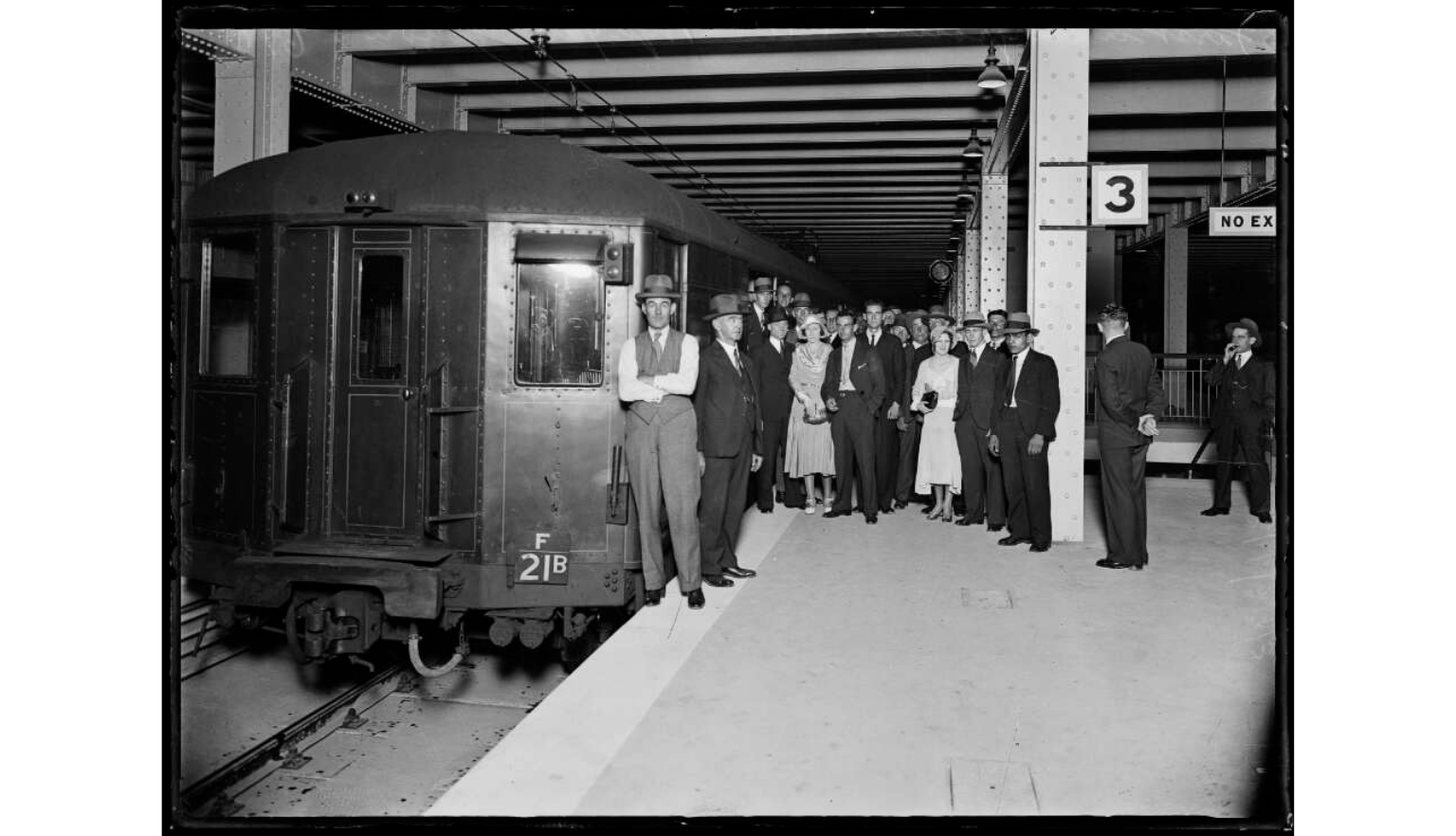 A group of men, including railway workers and officials, stand on a train platform next to a dark, vintage railway car inside a covered train station.