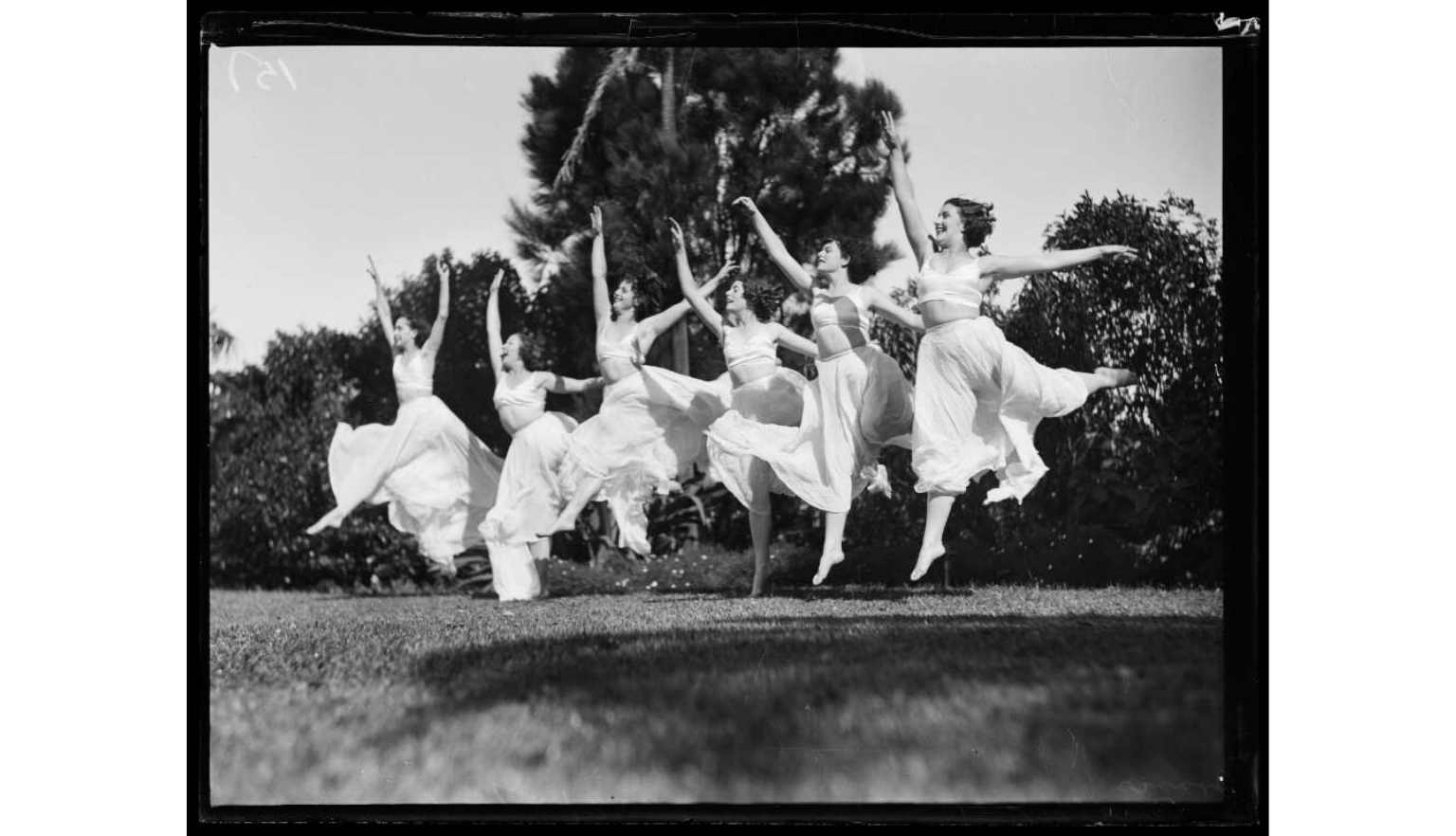 A group of female dancers in light-colored costumes are captured mid-air, performing an energetic routine outdoors in a grassy park setting under bright daylight.