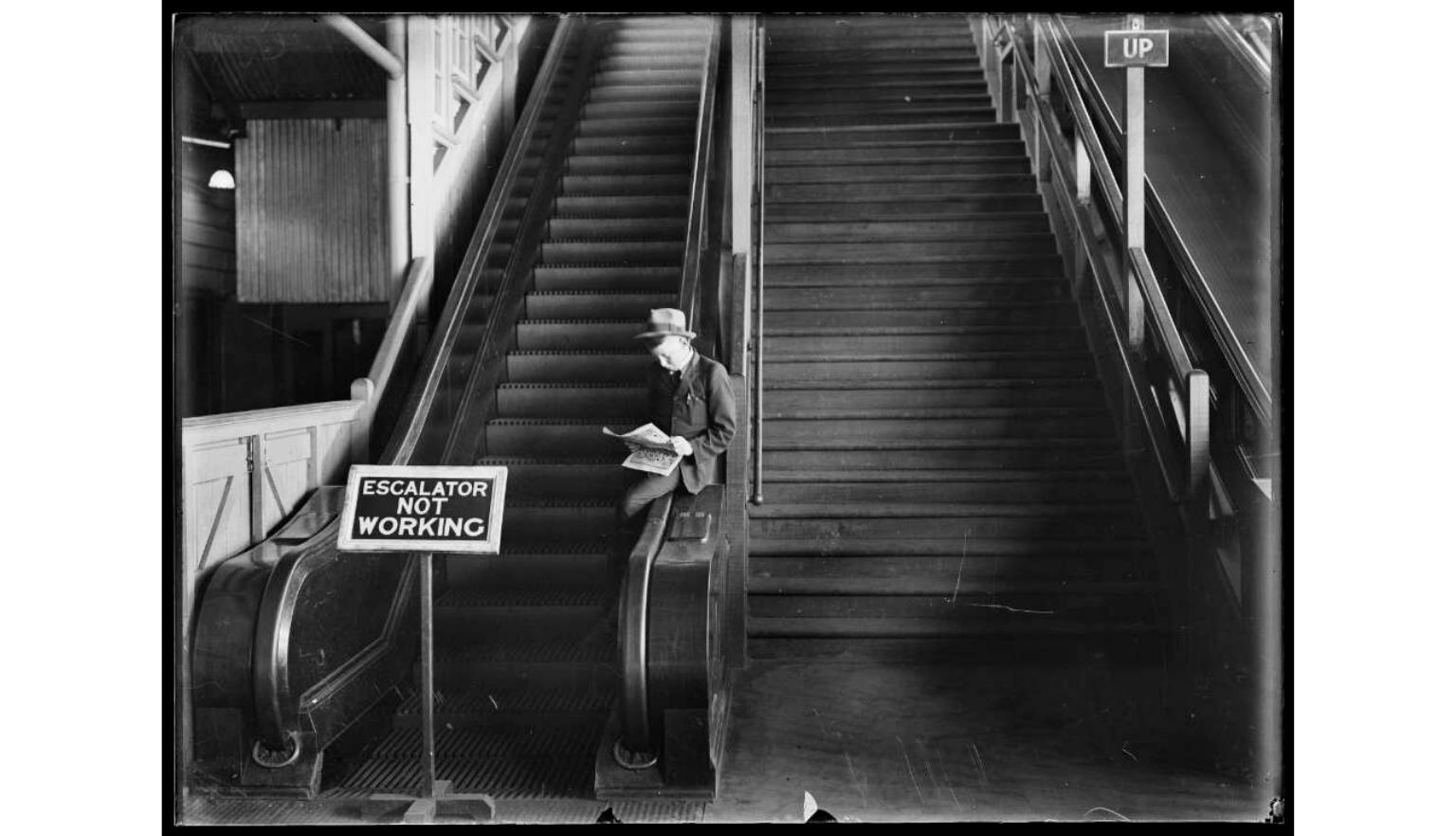 A black and white photograph showing an interior staircase area with a non-operational escalator. A man in a hat is seated on the steps, reading a newspaper.