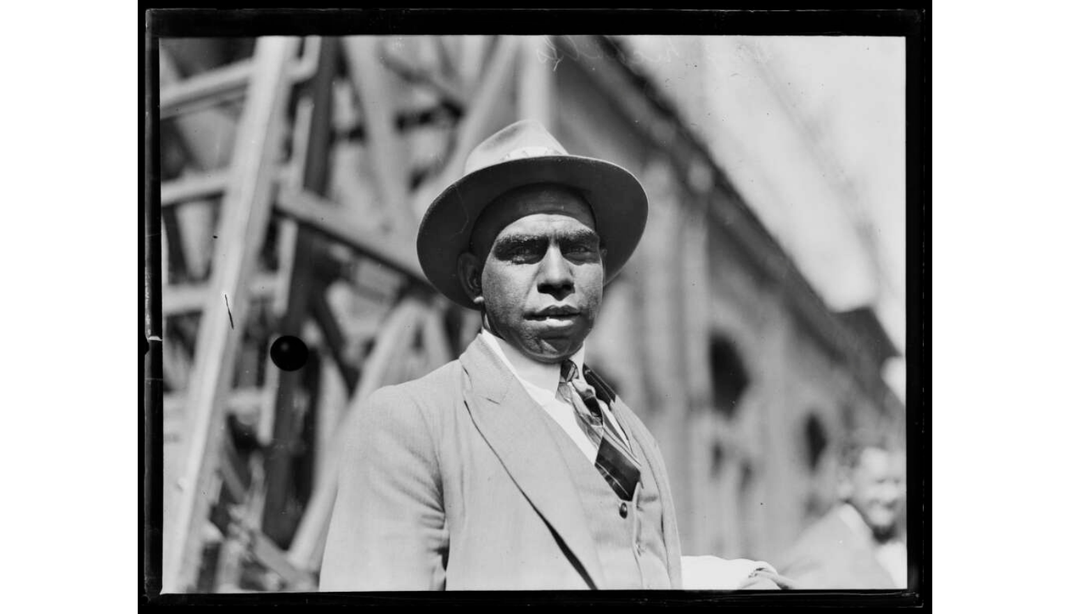 A black man wearing a hat and a light-colored suit jacket is photographed outdoors, looking directly at the camera. The background is somewhat blurred, showing industrial or urban structures.