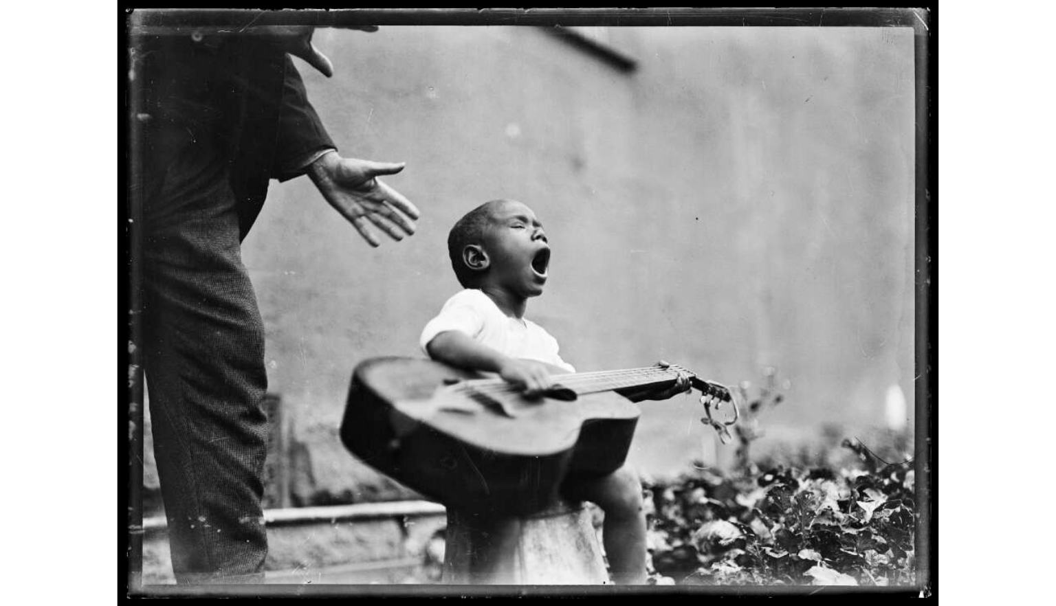 A black and white photograph captures a young child passionately singing while playing an acoustic guitar outdoors. An adult's arm and torso are visible in the upper left corner, seemingly interacting with the child.