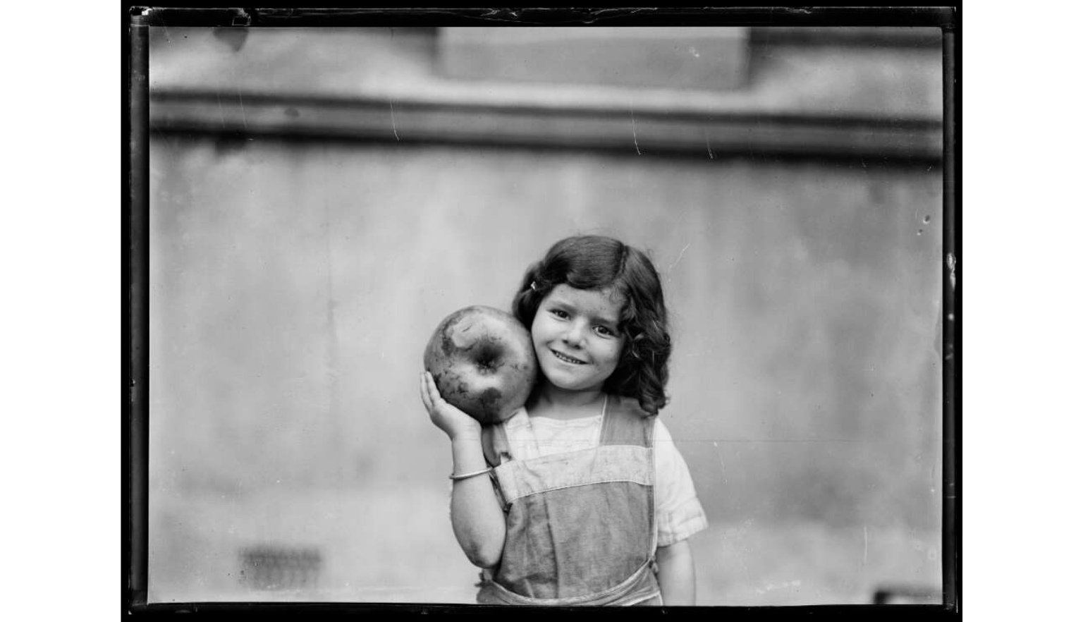 A black and white portrait of a young girl with curly hair, smiling while holding a large piece of fruit.