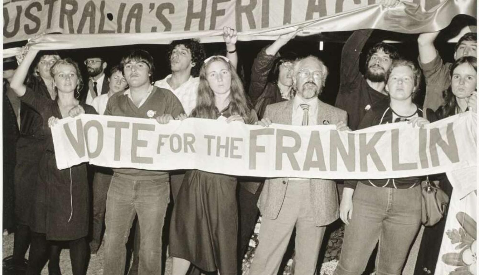 A black and white historical photo of a group of people marching and holding a large banner that reads "NOTE FOR THE FRANKLIN" under a larger banner that says "AUSTRALIA'S HERITAGE".