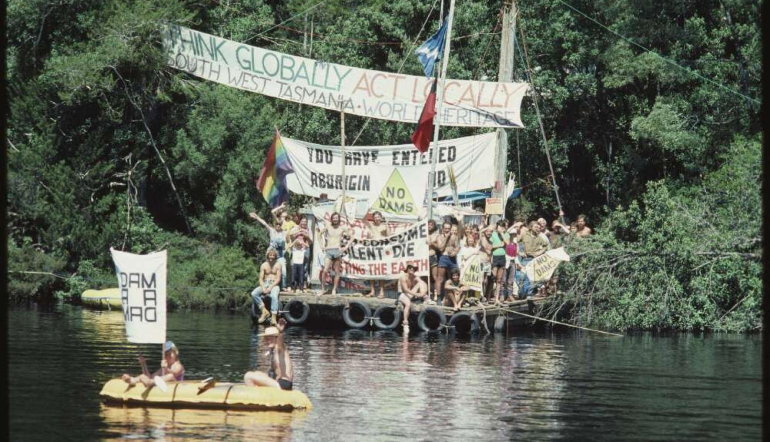 A group of people stands on a makeshift raft on a river, holding banners that read "THINK GLOBALLY ACT LOCALLY" and "YOU HAVE ENTERED ABORIGIN LAND".