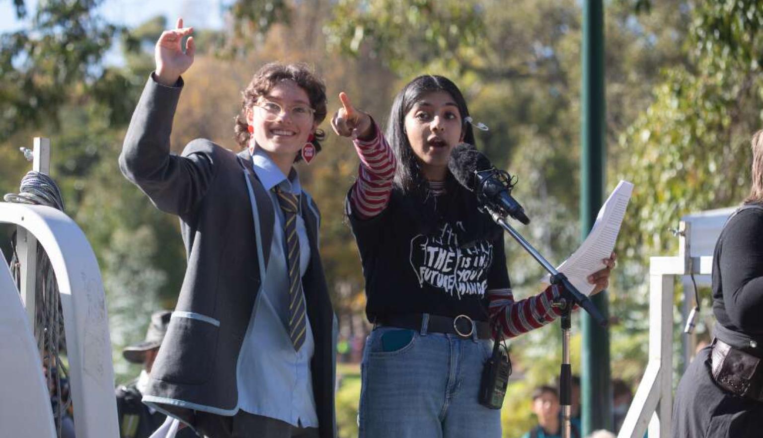 Two young students, enthusiastically speak outdoors. One of them holding a piece of paper. The other has their arm raised in a gesture of excitement.