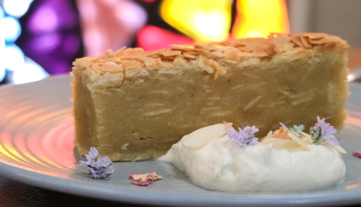 A dessert on a plate with cream and flowers with a colourful stained glass window behind it