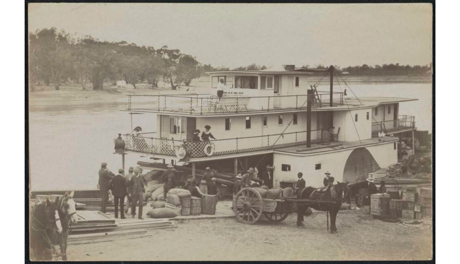 Historic paddle steamer docked at a riverside landing while workers load cargo from horse-drawn carts.