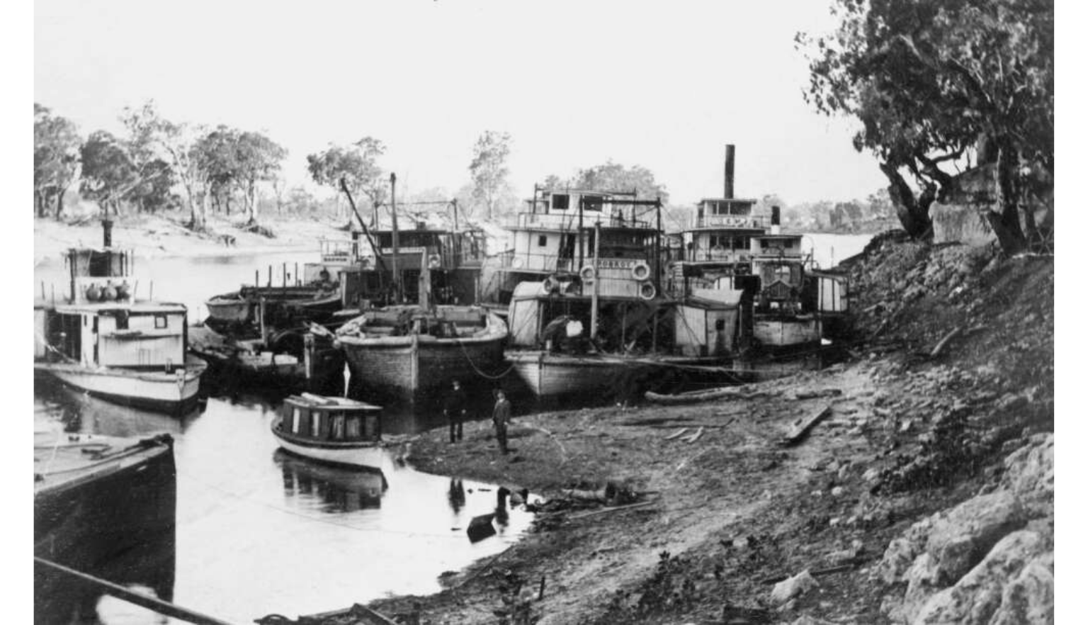 Several paddle steamers and smaller boats moored along a riverbank in a busy historic river port.
