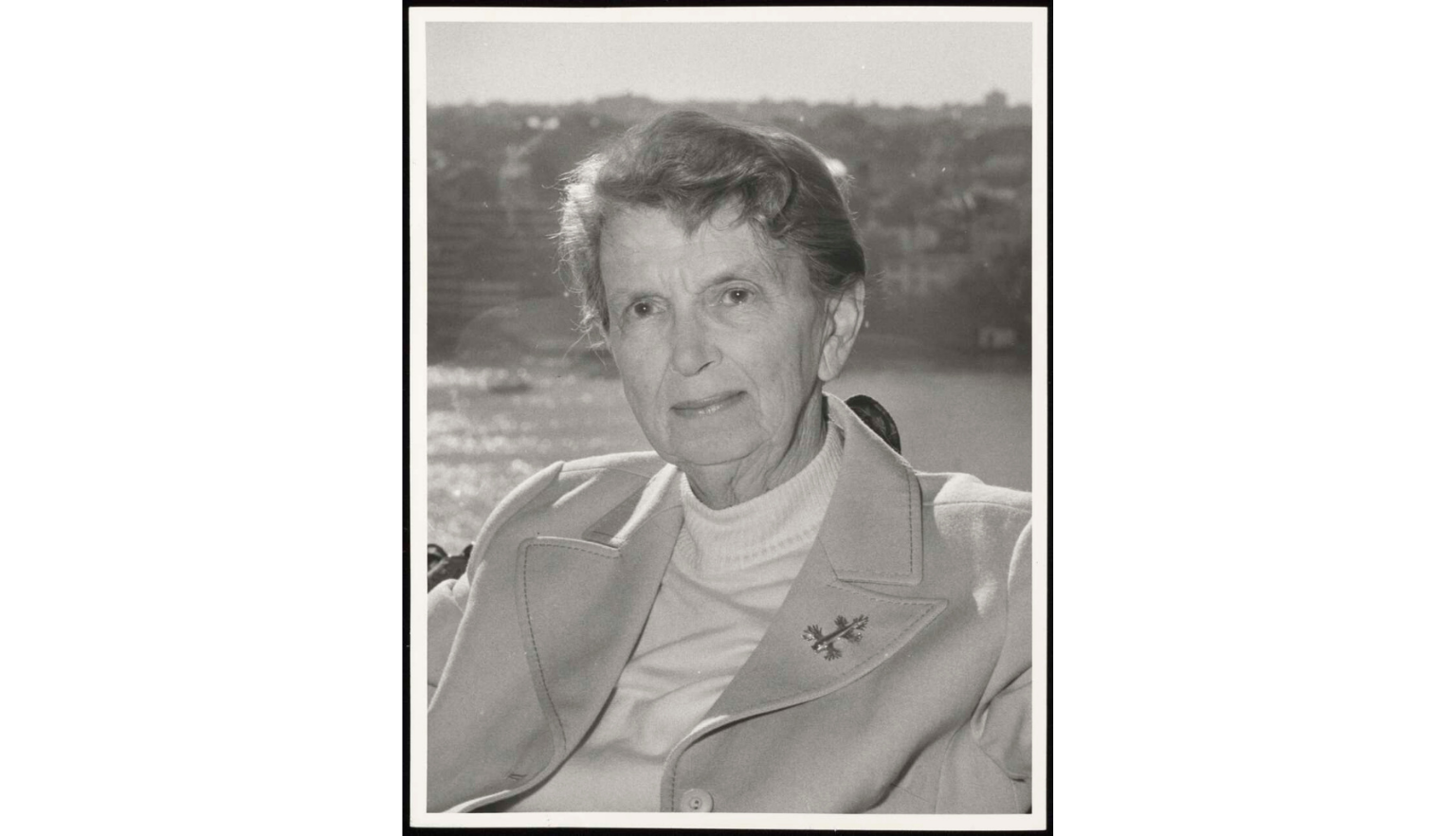 A black-and-white portrait of an older woman with short, styled hair, looking directly at the camera. She wears a light-colored coat over a turtleneck sweater, with a decorative brooch pinned to her lapel. The background is softly blurred, suggesting an outdoor setting with water and distant land.