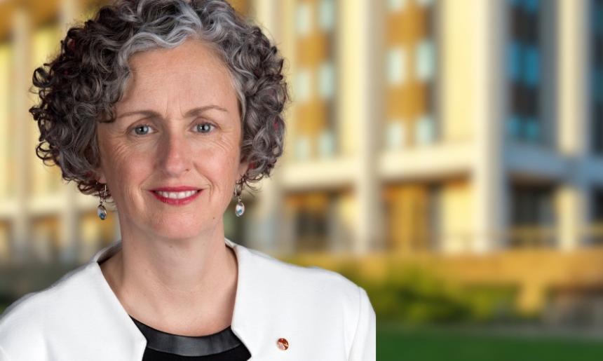 Woman with short, curly black and grey hair wearing a black top and white blazer standing in front of the National Library building