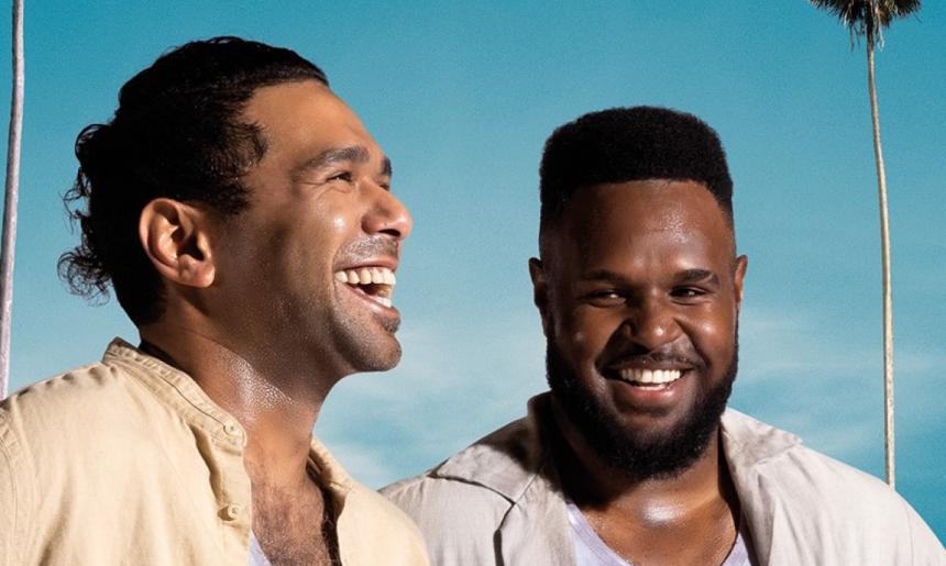 Two men smiling and laughing in front of a blue sky and palm tree.