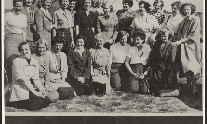 Black and white photo of 21 women of various ages smiling, standing in front of a plane. Below the image is a list of these women's names and where they're from