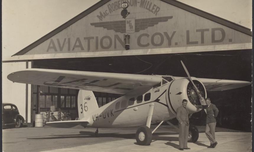 Two men standing and leaning against a small passenger plane in front of an aircraft hangar