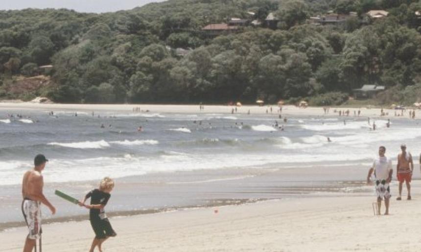 Group of men and a young boy playing cricket on the beach on a sunny, cloudless day.