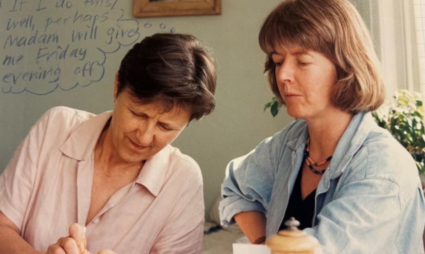 Two women looking down at something out of frame. One is wearing a pink shirt and one is wearing a blue shirt.