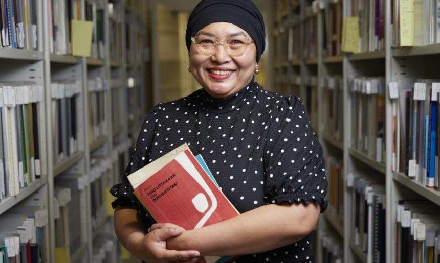 Woman wearing a dot print shirt, glasses and a black head scarf holding some booklets in an aisle of shelves