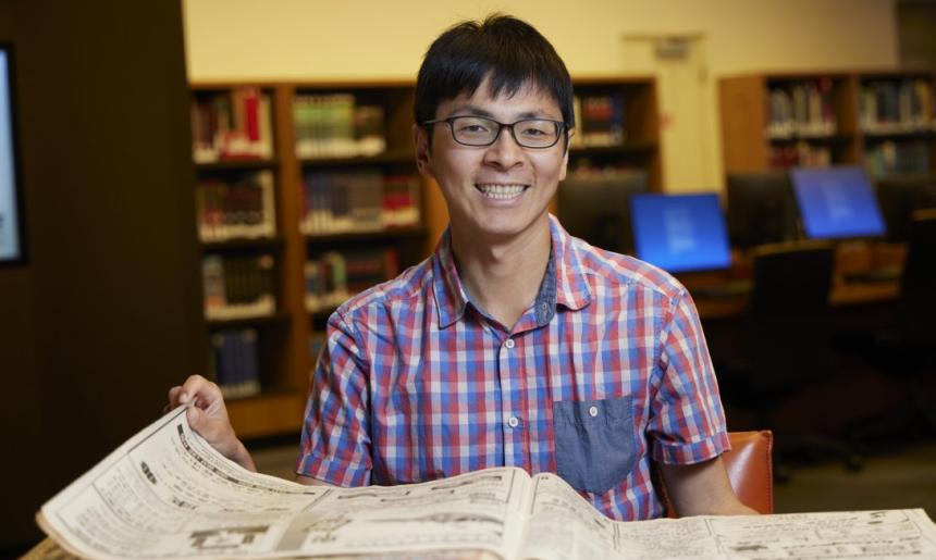 Man with short black hair and black glasses in a short-sleeved checkered shirt smiling and sitting at a desk with a Chinese language newspaper on it