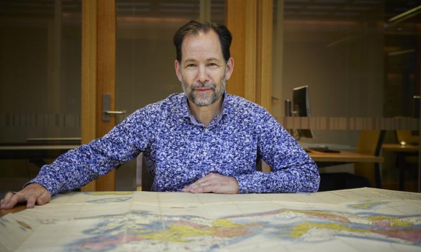 Man with brown hair and a grey and brown beard sitting at a table with a large map spread over it