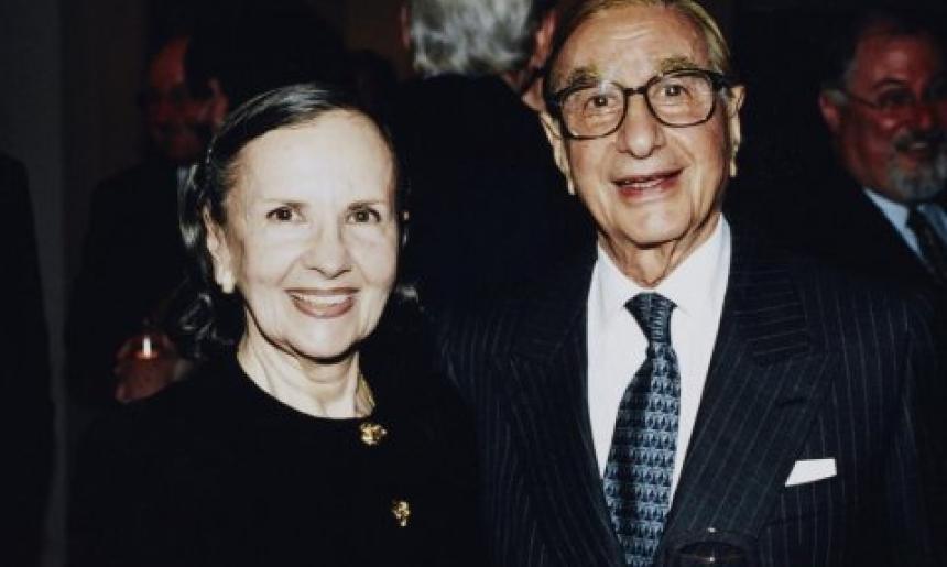 A couple smiles at the camera in a formal setting. Susan, with dark hair pulled back, wears a black outfit with gold buttons. Isaac, wearing glasses and a dark pinstriped suit, holds a wine glass in one hand. They are surrounded by a dimly lit background with other people.