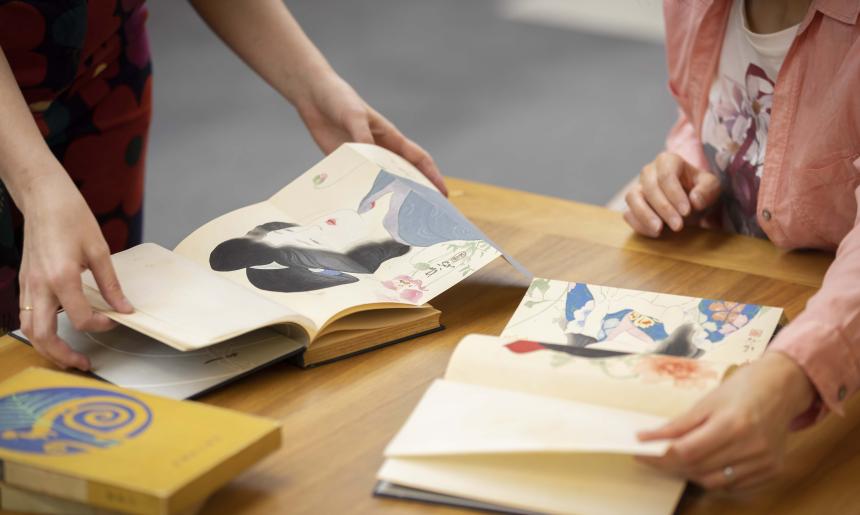 Two books open on a table with people flicking through them and looking at the books' colourful artwork showing drawings of women