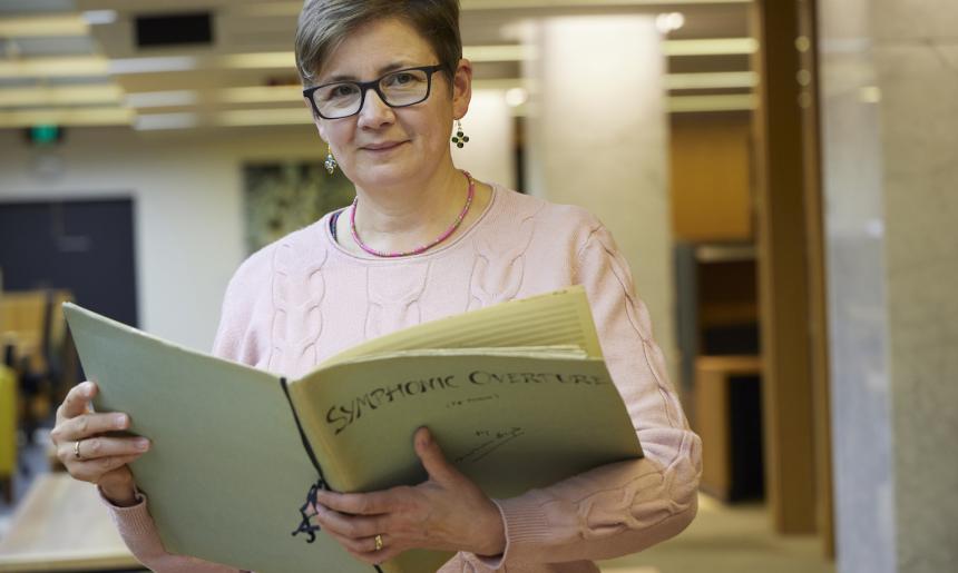 Celia Craig, a woman with very short brown hair, smiling and holding large book of sheet music with the title 'Symphonic Overture'