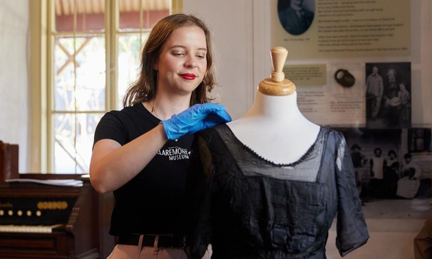 A person stands in Claremont Museum tending to a dress on a mannequin. There is a piano-style instrument, signage and a window in the background.