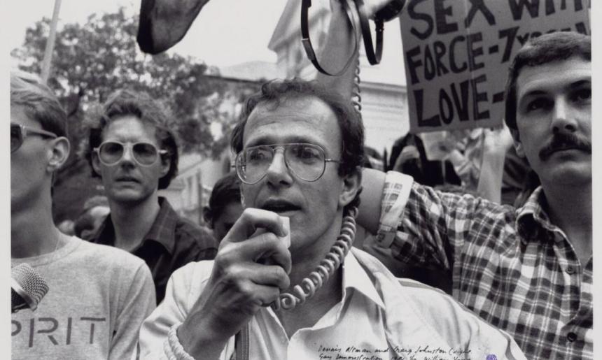 A black and white image of a group of mean demonstrating. The man front and centre is holding a microphone to his mouth, while the man to the right is holding a megaphone speaker up in the air. There are placards and flags in the air in the background.