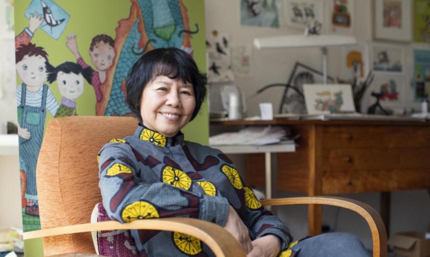A person sitting in a chair with their arm resting on the chair's arm. Behind them is a banner with the image of a dragon and three children on it. In the background is a desk and wall covered with illustrations and artworks.