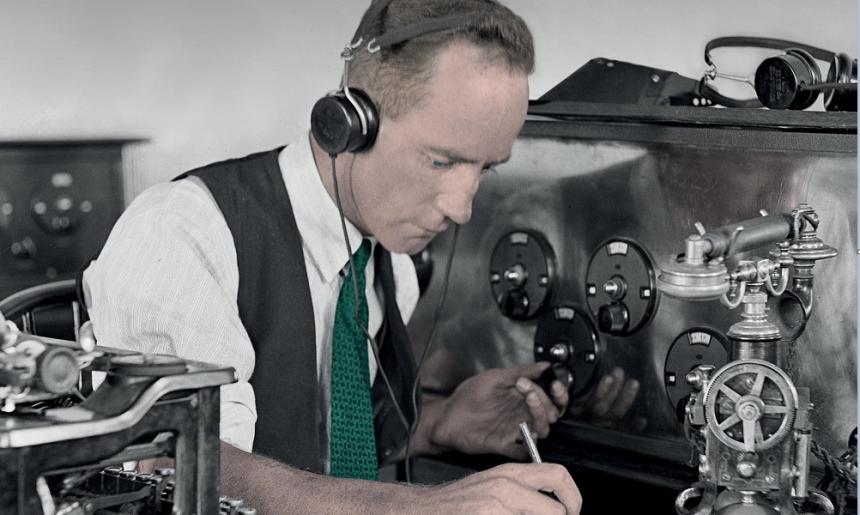 A man wearing old fashioned headphones is sitting at a table. He is writing on a notepad and is surrounded by lots of old fashioned communications equipment.