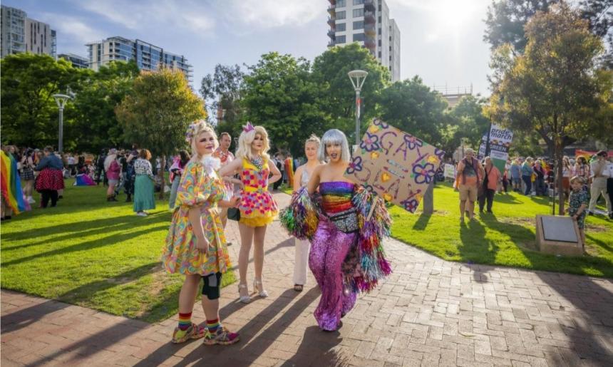 A group of four people who are very colourfully dressed, holding signs in a park. There are more groups of colourful people in the background.